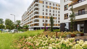 A modern multi-story building with balconies, a landscaped garden with hydrangea flowers, and a visible entrance with signage. - Modern Studio | Parking | Close to the Airport (Warszawa)