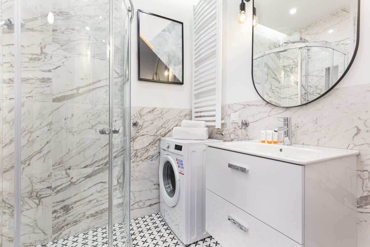 A modern bathroom with a glass shower enclosure, a built-in washing machine, a white vanity with a mirror, and a marble-tiled floor.