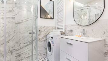 A modern bathroom with a glass shower enclosure, a built-in washing machine, a white vanity with a mirror, and a marble-tiled floor.