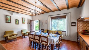Comedor con mesa de madera dispuesta para cuatro personas, rodeada de sillas de madera. Se ve una ventana con cortinas, una araña y una zona de cocina con salpicadero de azulejos.
