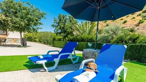 Dos sillones azules con estructura blanca, un paraguas grande y una mesa de piedra con un sombrero y un libro.