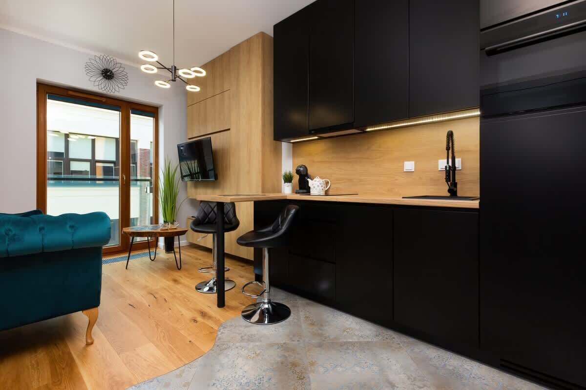 A modern kitchen with a wooden countertop, black cabinets, and a black refrigerator. There is a bar stool and a teal sofa in the living area.