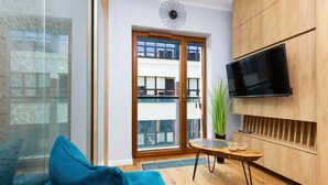 A modern living room with a blue sofa, a wooden cabinet, a flat-screen TV, and a large window with a view of buildings. - Modern Studio in the Center | Balcony | Close to Attractions (Warszawa)