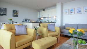 Una sala de estar moderna con sofás amarillos, una mesa de centro de vidrio y una cocina al fondo.