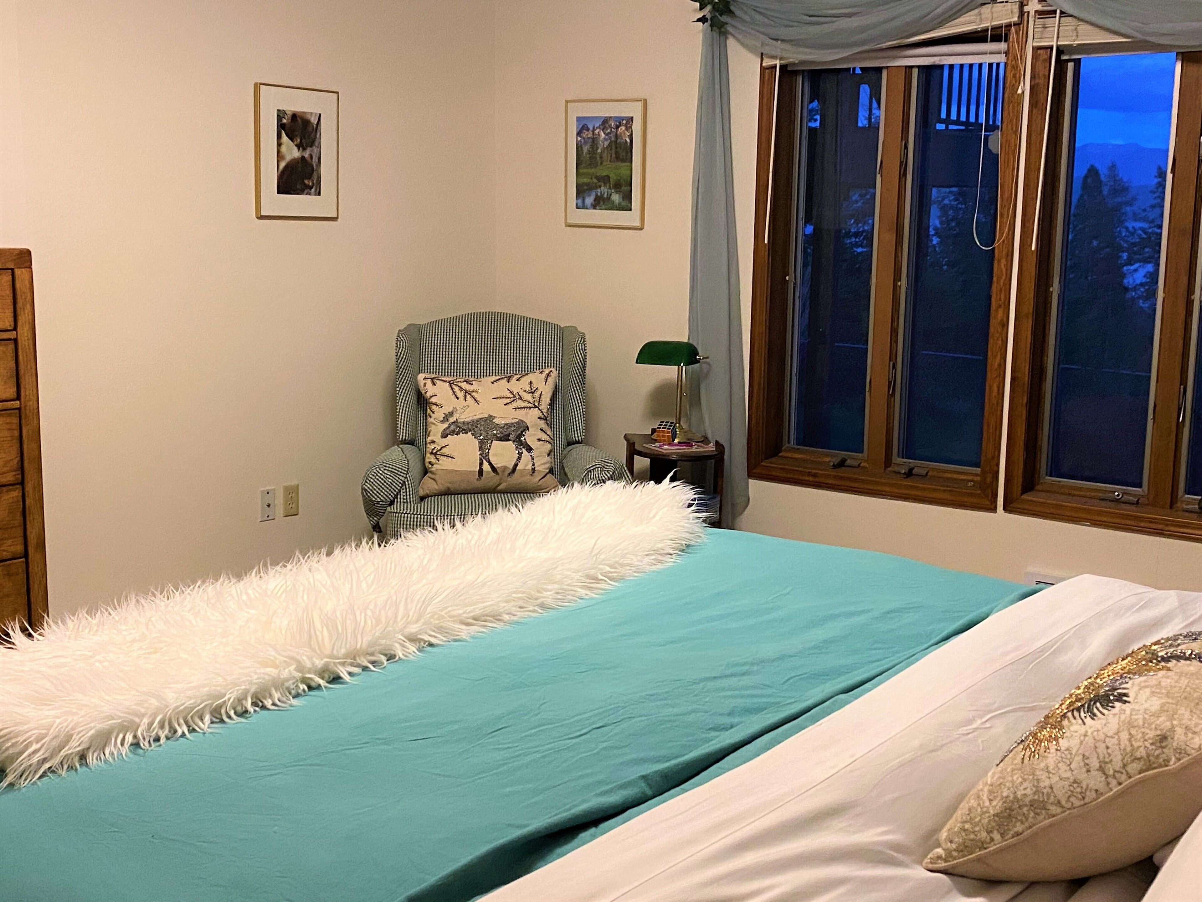 A bedroom with a bed, a chair with a moose pillow, a nightstand, and a window with a view of trees.