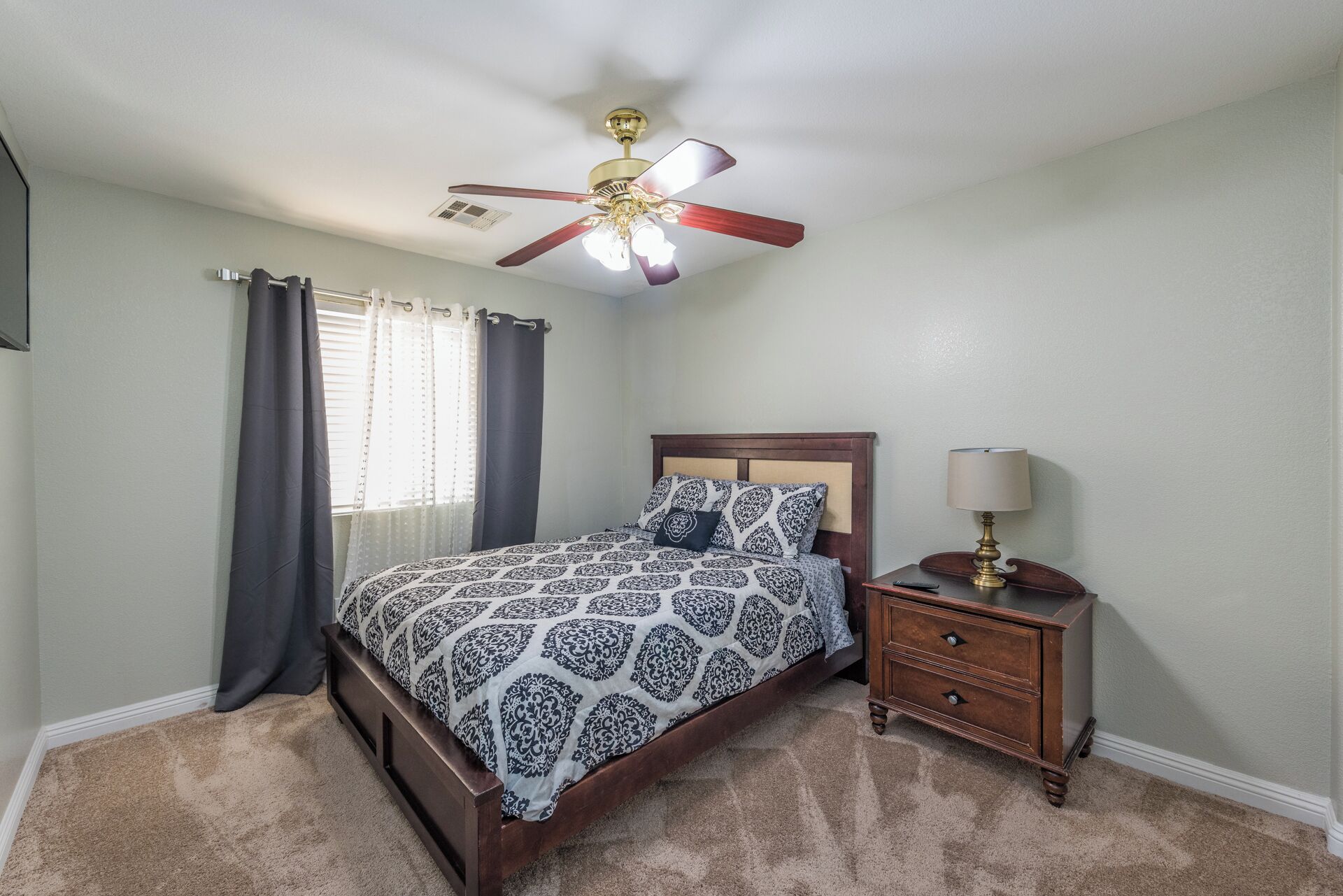 A bedroom with a bed, a nightstand, a lamp, a ceiling fan, and a window with curtains.