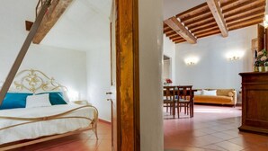 Quarto com cama, porta de madeira e vista para uma sala de estar com sofá e mesa de jantar.
