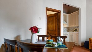 Uma sala de jantar com uma mesa de madeira, cadeiras, um vaso com flores e uma cozinha ao fundo.