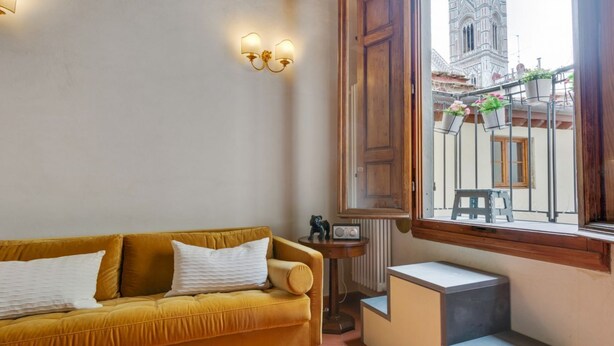 Um quarto com um sofá cor de mostarda, uma mesa pequena com um rádio e uma janela com vista para um edifício histórico.