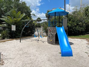 Une aire de jeux avec un toboggan bleu et des balançoires, entourée d’arbres et recouverte de sable.