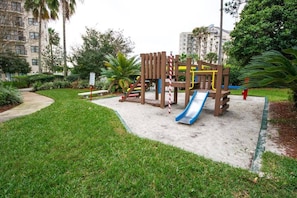 Children can play safely on the playground's slide and climbing elements surrounded by lush greenery and trees.