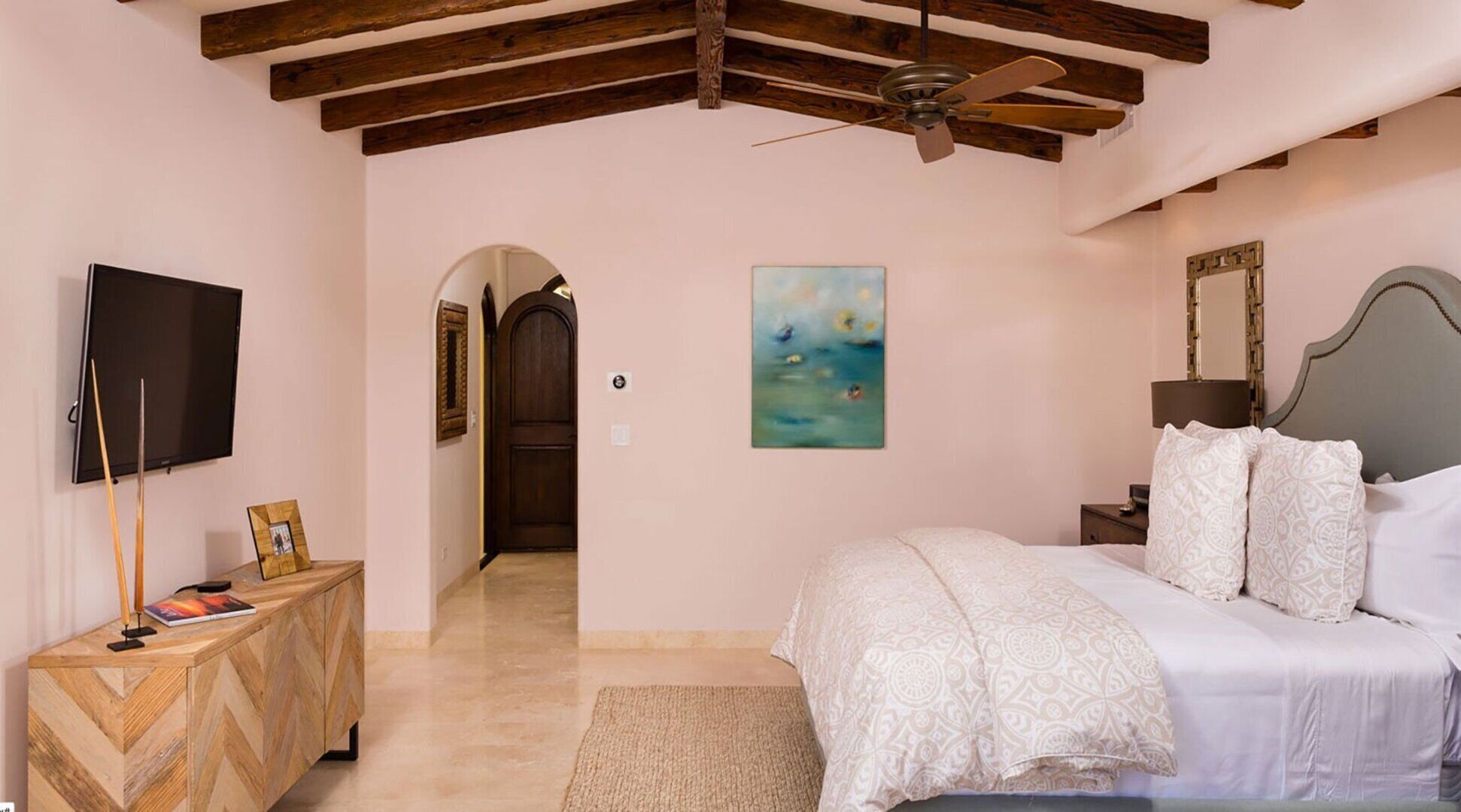 A bedroom with a bed, a TV, a ceiling fan, a painting, and a wooden cabinet.