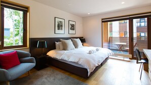 A modern bedroom with a large bed, a grey sofa, a small table, and a balcony with wooden blinds. - Exclusive 5 Bedroom Luxury Chalet, Niseko Chalet 1004 (Niseko)
