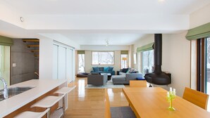 Una cocina moderna con una isla central, un comedor con mesa y sillas, una sala de estar con sofás y chimenea, y ventanas grandes con vistas a árboles.