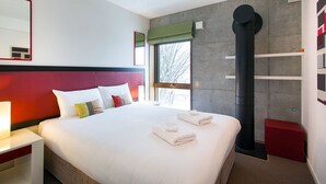 Una habitación de hotel moderna con una cama grande, una mesita de noche con lámpara, un ventanal con vistas y una estructura de columna negra distintiva.