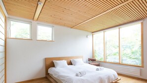A modern bedroom with a wooden slatted ceiling, a bed with white linens, and large windows offering a view of greenery.