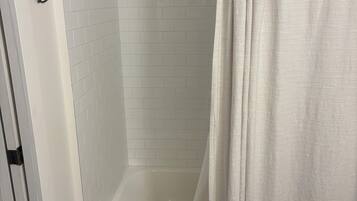 Un baño con pared de ducha en blanco y azulejos, una bañera blanca y suelo de madera.
