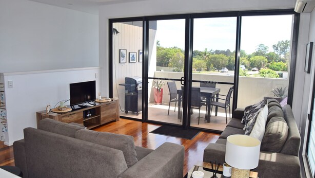 Ein modernes Wohnzimmer mit Flachbildfernseher, einer Couch, einem Essbereich mit Stühlen und einem Balkon mit Grill und Topfpflanze.
