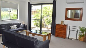 Ein Wohnzimmer mit Sofa, Couchtisch und Kommode. Ein Glastür öffnet sich zu einem Außenbereich mit Pflanzen.