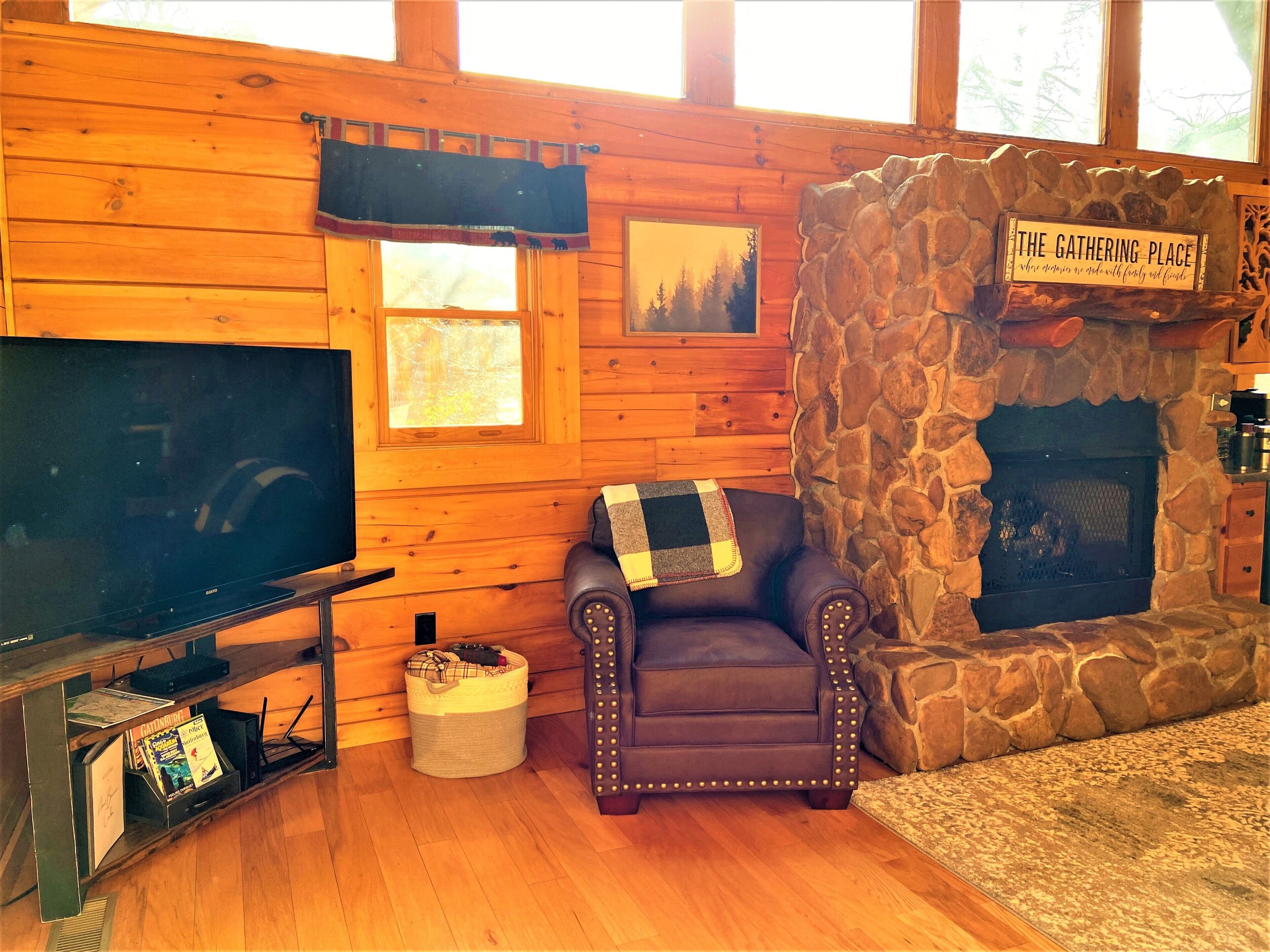 A cozy cabin interior with a stone fireplace, a leather armchair, a television, and a sign reading 'The Gathering Place'.