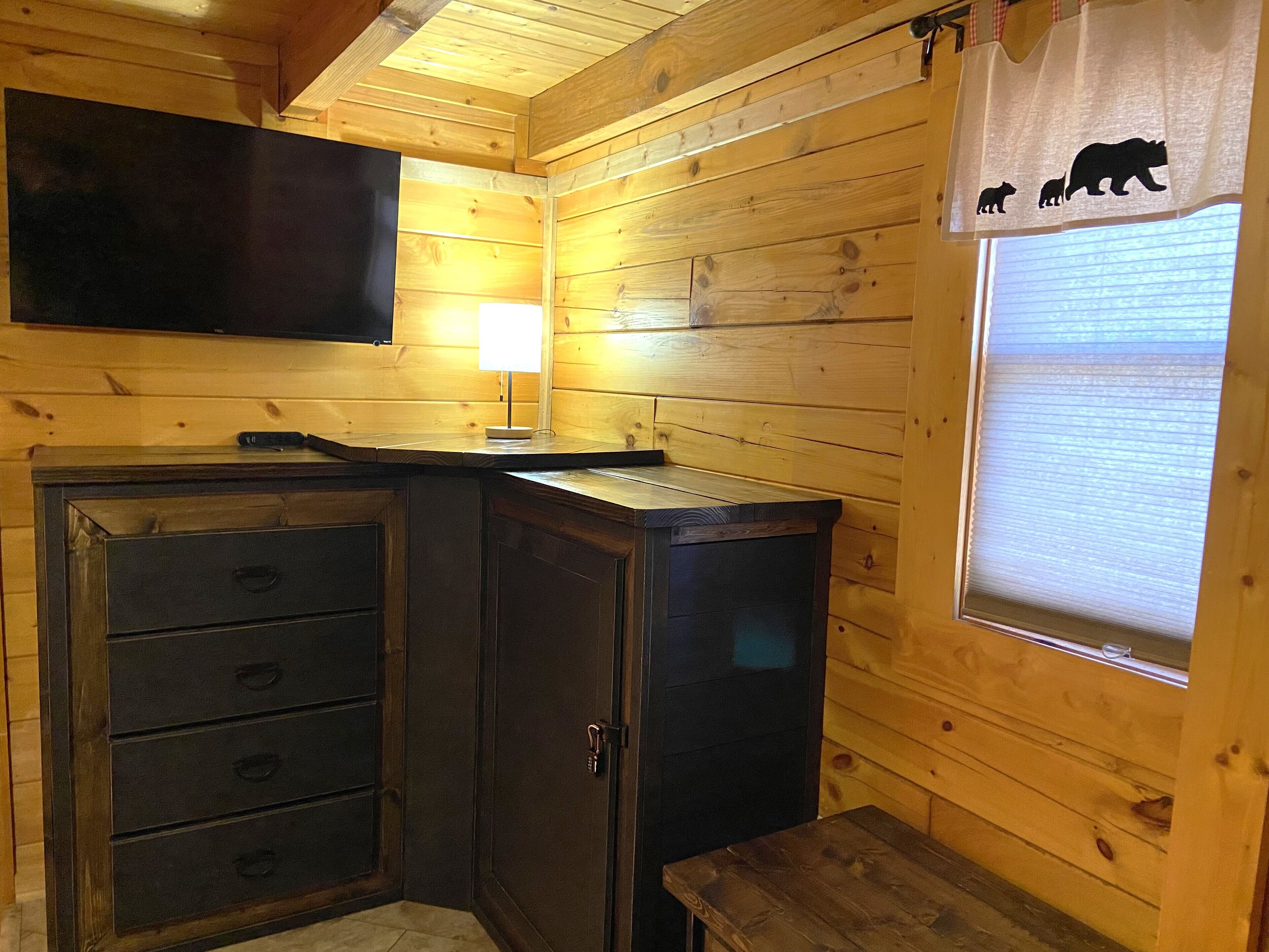 A wooden cabin interior with a TV mounted on the wall, a desk with a lamp, and a window with a bear-themed curtain.