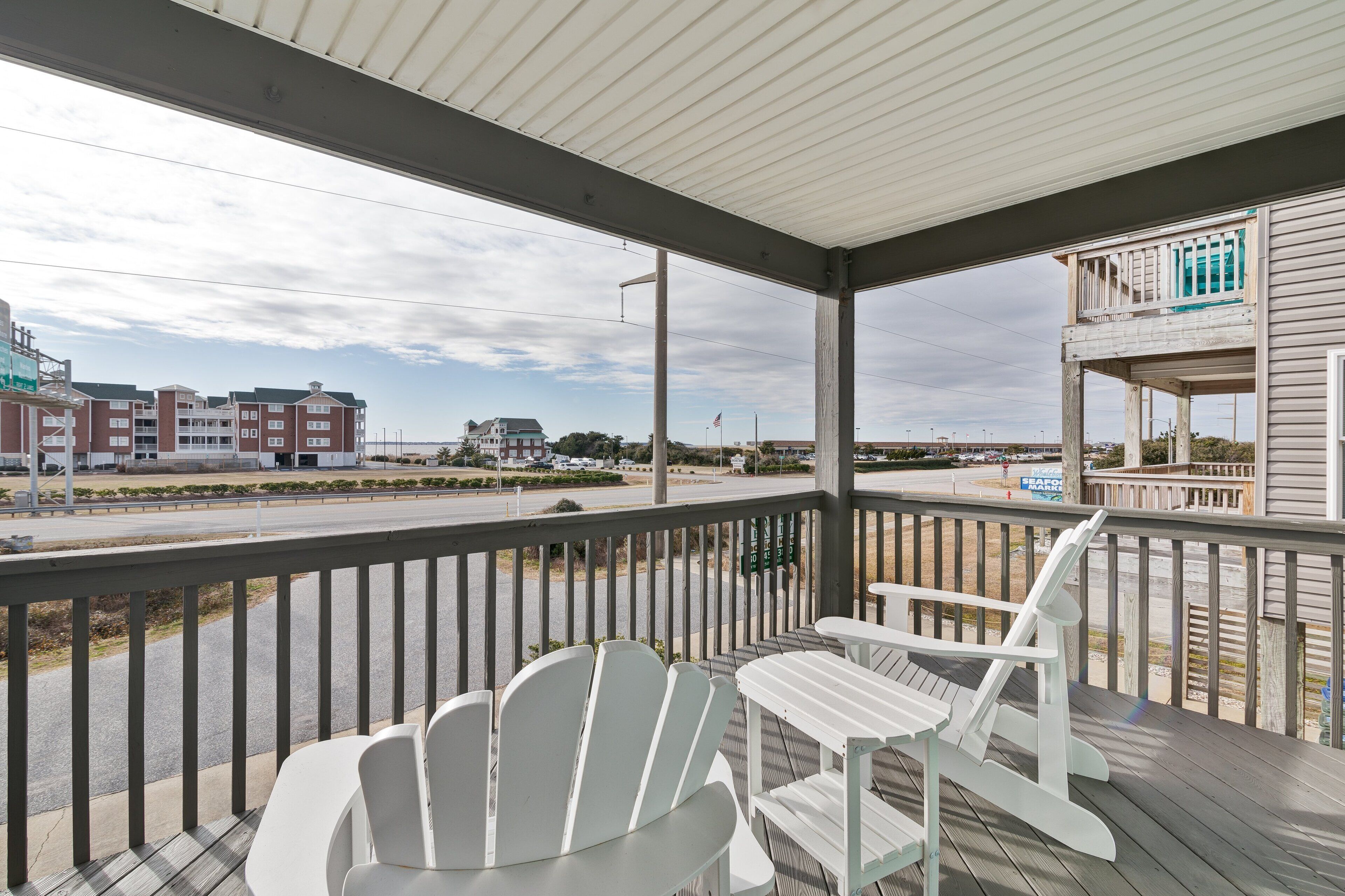 A covered porch with white Adirondack chairs, a view of a road, and a building with a balcony.