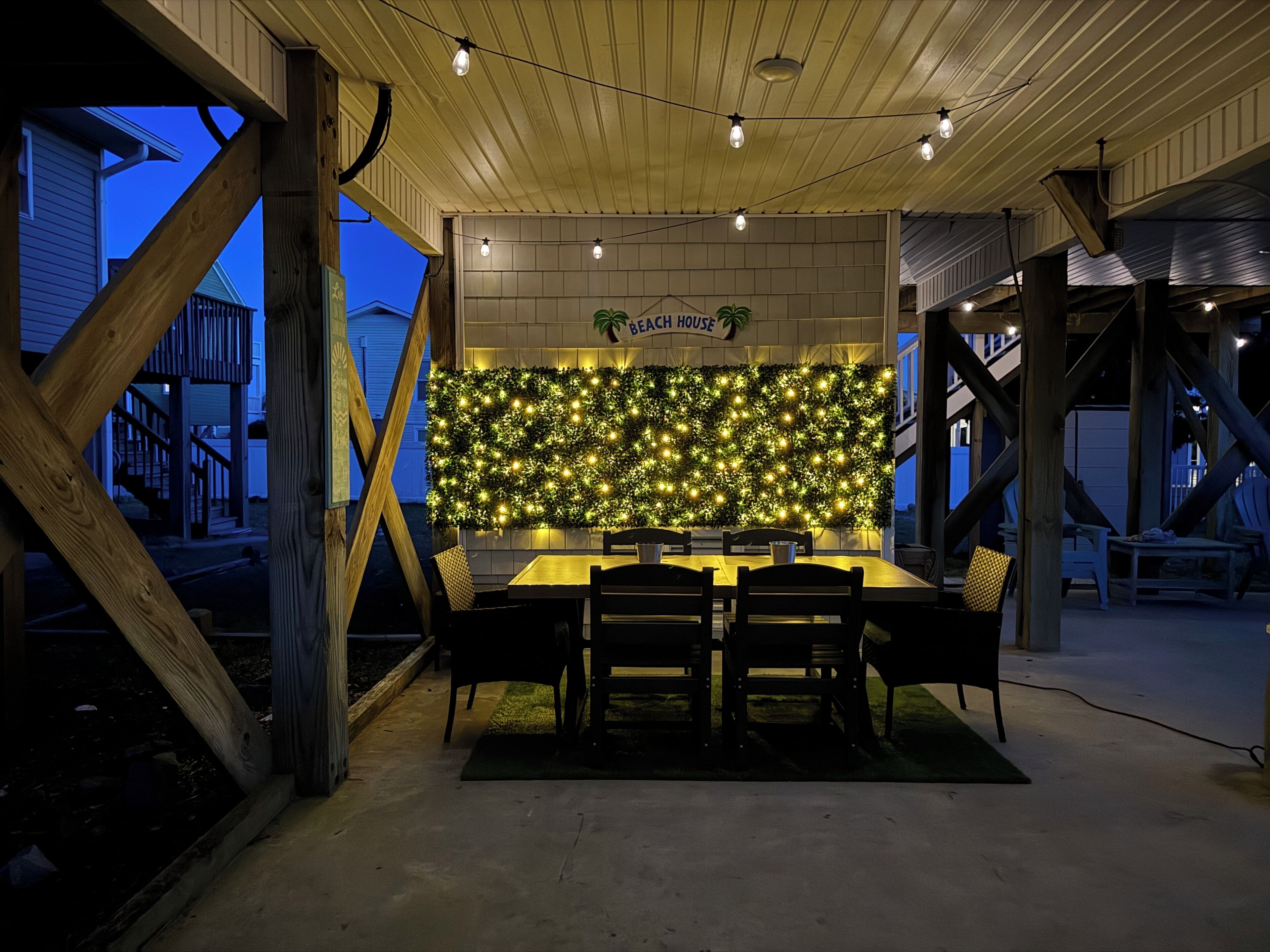 Uma área de refeições ao ar livre com uma mesa e cadeiras de madeira, uma parede decorada com luzes, e uma placa com a inscrição 'Beach House'.