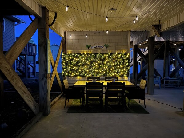 An outdoor dining area with a wooden table and chairs, a wall decorated with lights, and a sign that reads 'Beach House.'