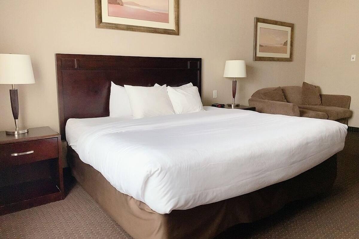 A hotel room with a large bed, two bedside tables with lamps, a brown armchair, and two framed pictures on the wall.
