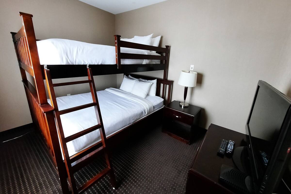A hotel room with a bunk bed, a desk with a lamp, a television, and a small drawer.