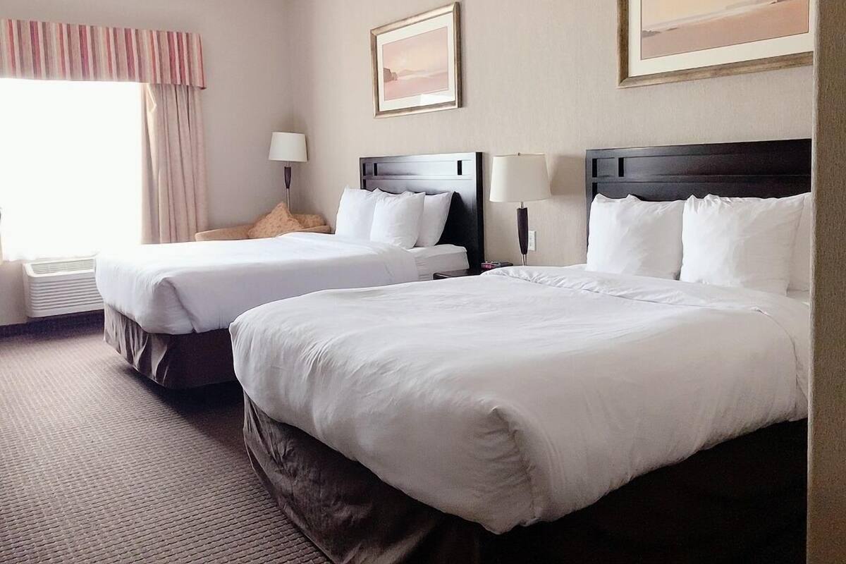 A hotel room with two beds, a window with curtains, a lamp, and two framed pictures on the wall.