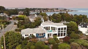 Une maison moderne à deux étages, dotée d’un grand balcon, entourée d’une végétation luxuriante et offrant une vue sur la mer.