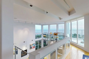A spacious interior with large windows offering a view of the sea, a balcony with outdoor seating, and a modern staircase.