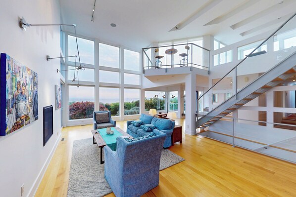 A spacious living area with a high ceiling, large windows, a staircase, and modern furniture.