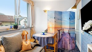 Una habitación con sofá, una mesa redonda con mantel azul, una silla, una estructura decorativa de madera y vistas al atardecer a través de una ventana.