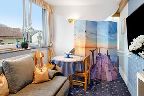A room with a sofa, a round table with a blue tablecloth, a chair, a decorative wooden structure, and a view of the sunset through a window.