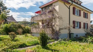 Una casa de dos pisos con un balcón, rodeada por un jardín con diversas plantas y flores.
