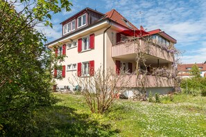 A two-story building with a balcony, surrounded by greenery and white flowers.