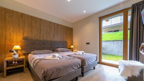 Un dormitorio con una cama grande, dos mesitas de noche, un reposapiés blanco y mullido, y un ventanal corredero que da a un balcón.