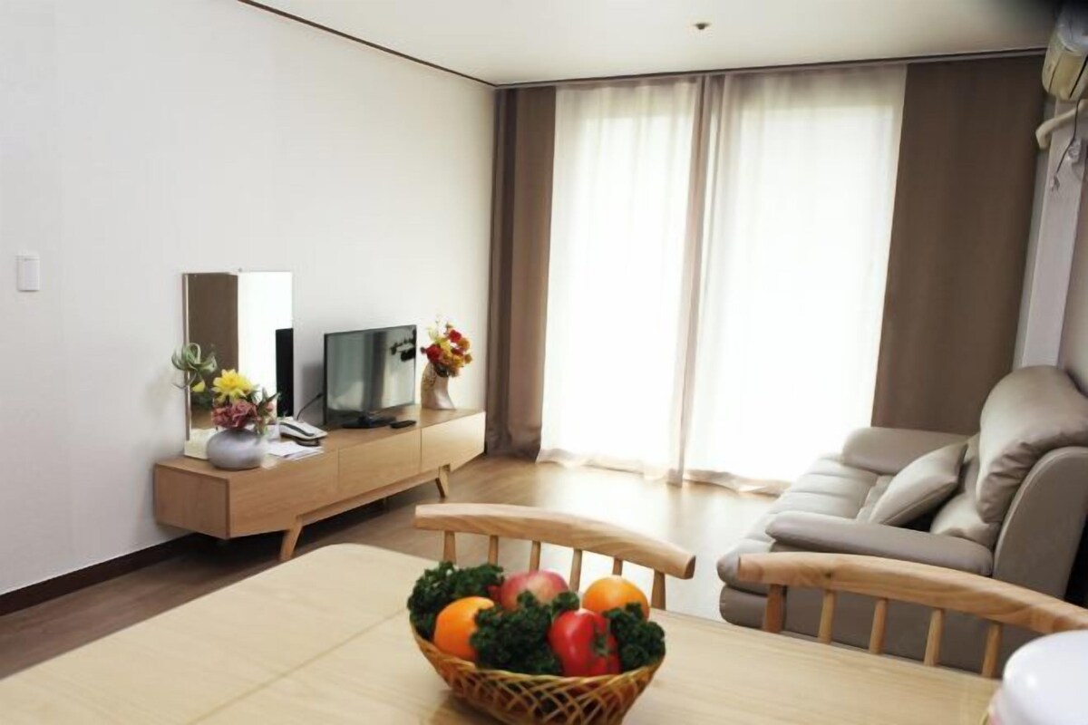 Una sala de estar moderna con un sofá, un televisor sobre un mueble de madera, una mesa de comedor con una cesta de frutas y un ventanal con cortinas.