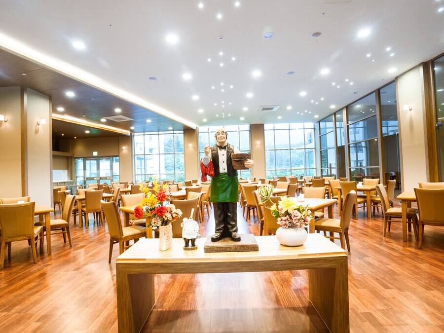 A spacious dining area with wooden tables and chairs, a central statue, and large windows allowing natural light.
