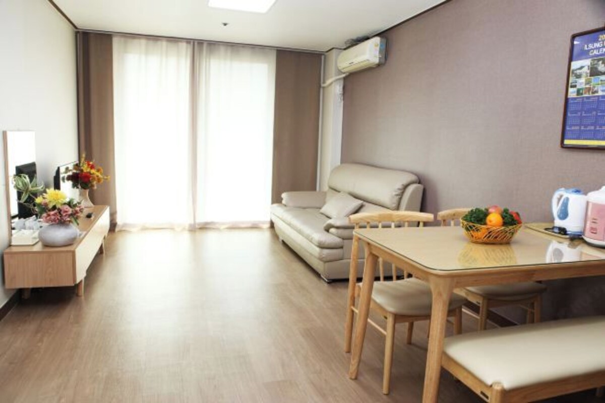 A modern hotel room with a sofa, a dining table with chairs, a small kitchen area, and a large window with curtains.