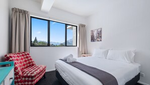 A bedroom with a bed, a chair, a window with a view, and a painting on the wall. - Brookside Gem Modern 2 Bedroom Apartment (Queenstown)