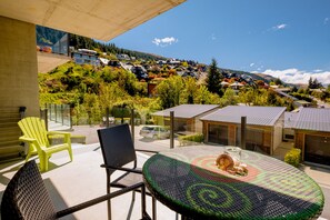A balcony with wicker chairs, a glass table, and a view of a hillside with houses. - Brookside Gem Modern 2 Bedroom Apartment (Queenstown)