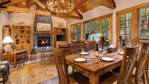 A spacious room with a large wooden dining table, stone fireplace, and large windows offering a view of the outdoors. - Abode at the Homestead, walk to slopes, hot tub (Teton Village)