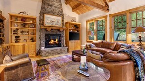 A cozy living room with a stone fireplace, leather furniture, and a large window offering a view of the outdoors. - Abode at the Homestead, walk to slopes, hot tub (Teton Village)