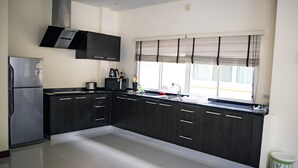 A modern kitchen with dark cabinetry, a stainless steel refrigerator, and a window with blinds. - Villa Tanya at Platinum (Rawai)
