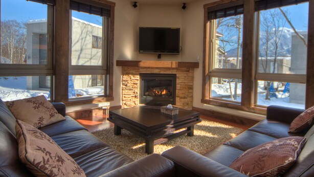 A cozy living room with a stone fireplace, leather sofas, and large windows offering a view of a snowy landscape.