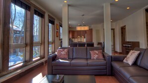 A spacious living room with a large brown leather sofa, a coffee table, and a view of a kitchen and staircase.