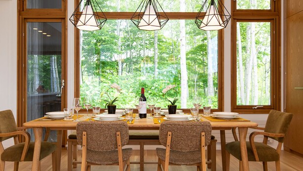 A dining area with a table set for six, surrounded by chairs, with a view of trees through large windows. - Luxury 5 Star 3 Bedroom Chalet, Niseko Chalet 1005 (Niseko)